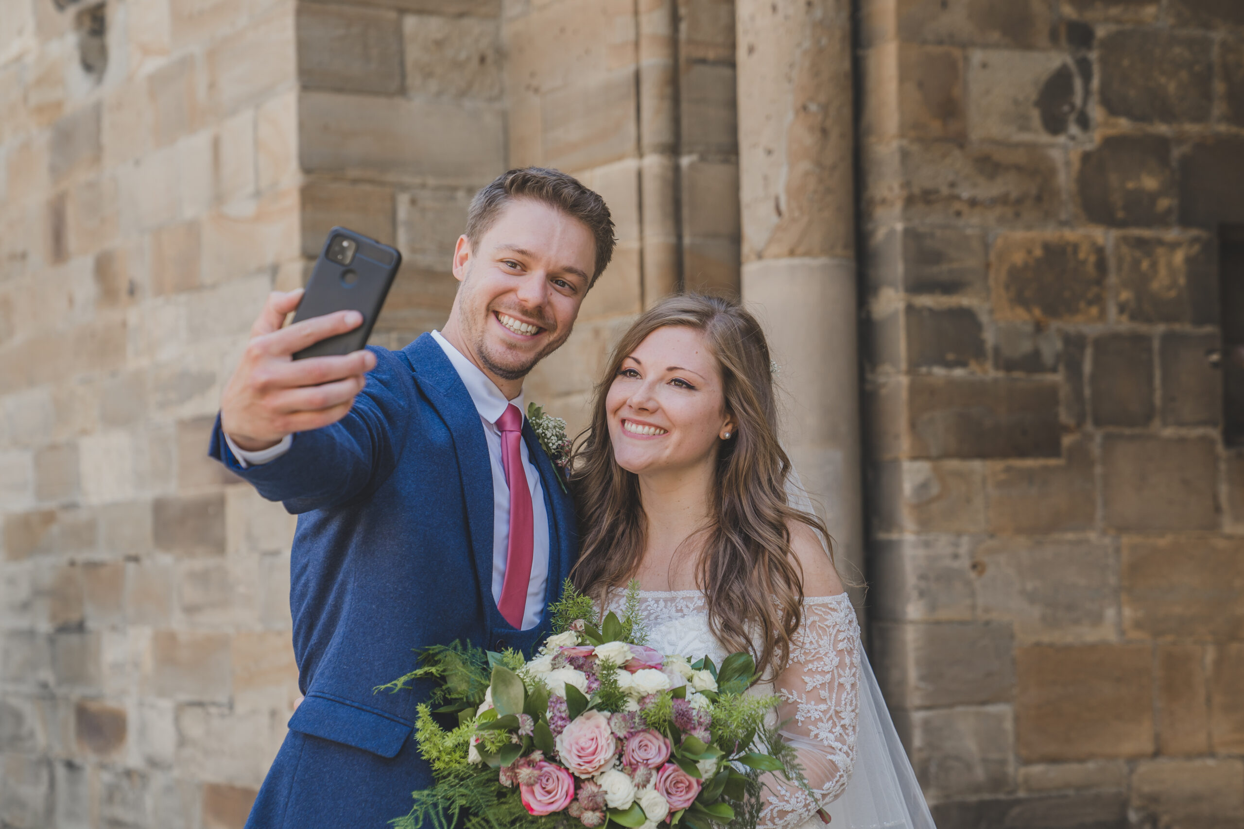 A bride and groom taking a selfie in front of a stone building.