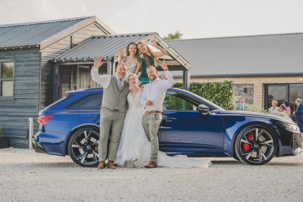 The bride and groom pose with their wedding car in front of a barn.