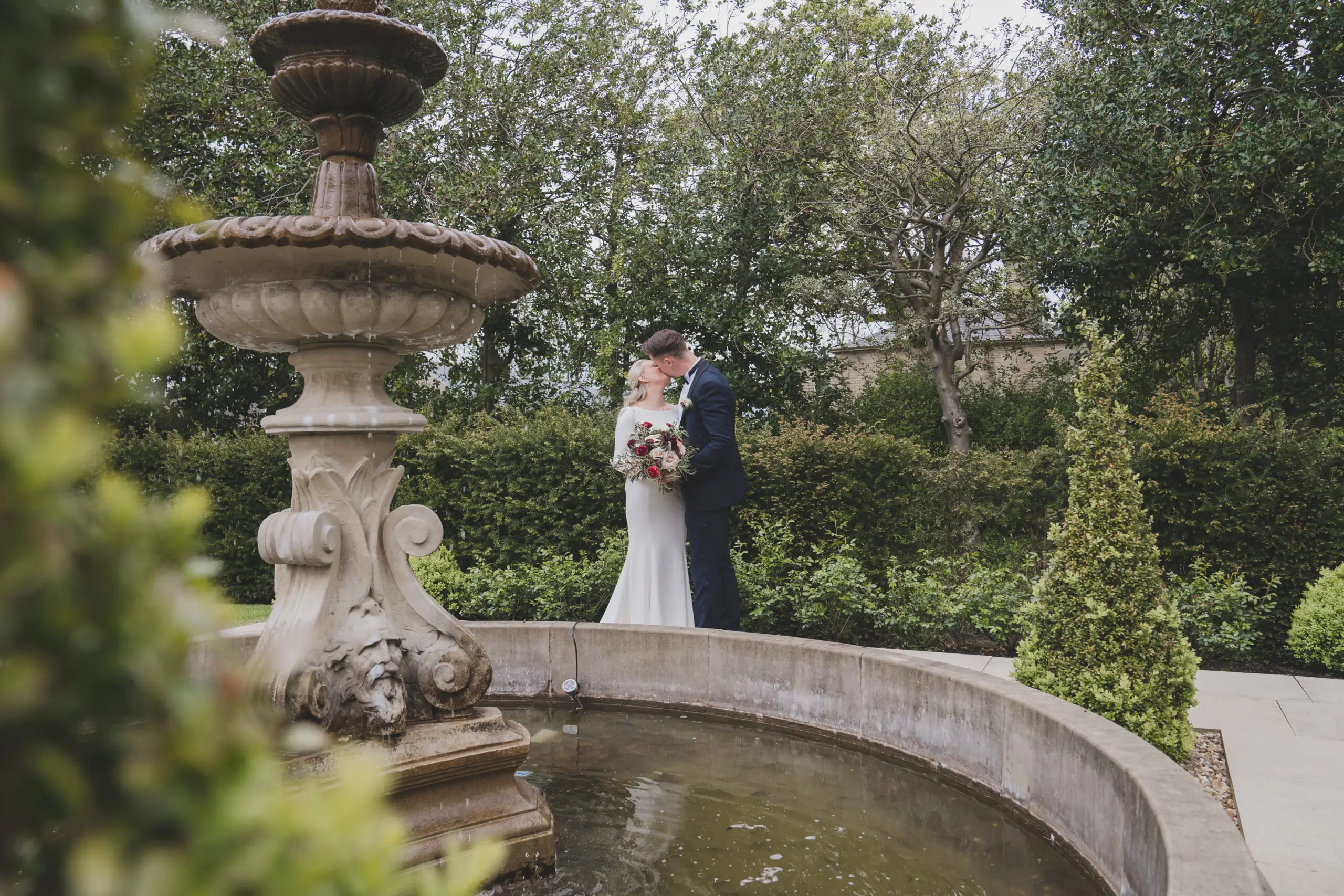 A bride and groom share a kiss next to an ornate stone fountain surrounded by lush greenery in a garden setting.
