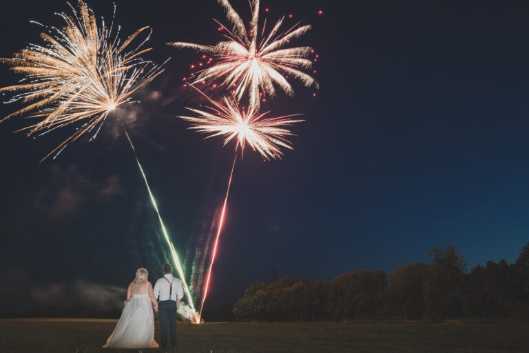 A newlywed couple stands in a field at night, watching colorful fireworks explode in the sky above.